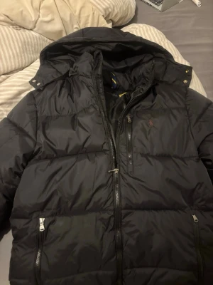 Svart dunjacka från Polo Ralph Lauren - Snygg svart dunjacka från Polo Ralph Lauren med huva och dragkedja framtill. Jackan har tre fickor med dragkedja och den klassiska Polo-loggan broderad på bröstet. Perfekt för kalla vinterdagar och riktigt skön att bära.