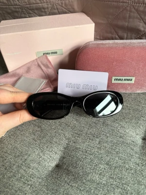 Svarta solglasögon från Miu Miu - Snygga svarta solglasögon från Miu Miu med breda bågar och guldiga detaljer på sidan där loggan står ut. Glasögonen har en klassisk, lite kantig form och kommer med originalfodral i rosa sammet.