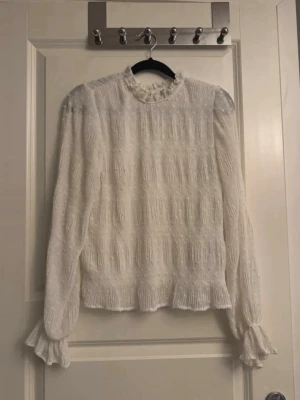 Vit volangblus från Nelly - Söt vit blus från Nelly med volangdetaljer vid hals och ärmslut. Blusen har långärmad design, diskret strukturmönster och små prickar i tyget. Perfekt för dig som gillar romantisk och trendig stil.