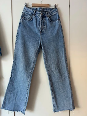 Blå raka jeans med rå kant - Säljer ett par ljusblå raka jeans med hög midja och råa, avklippta benslut. Jeansen har klassiska fem fickor och stängs med knapp och dragkedja. Perfekta för en avslappnad och trendig look.