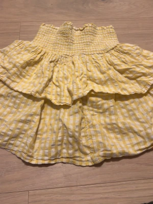 Gul och vit rutig volangkjol - Supersöt kortkjol i gul och vit rutig bomull med bred smock i midjan och dubbla volanger. Perfekt för varma dagar och ger en riktigt somrig vibe. Lätt och luftig modell som är enkel att matcha med en enkel topp.