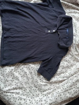 Mörkblå pikétröja från Gant - Klassisk mörkblå pikétröja från Gant med korta ärmar och ribbade muddar. Tröjan har två knappar vid halsen och en mjuk krage. Perfekt till jeans eller shorts för en clean och enkel stil.