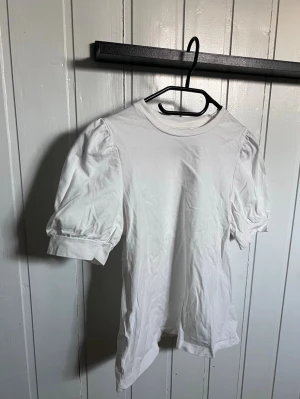 Vit puffärmad topp i bomull - Säljer en vit topp med puffärmar och rund halsringning. Toppen har en enkel och stilren design med korta ärmar som ger en trendig siluett. Tillverkad i mjuk bomull som känns skön mot huden. Perfekt att matcha med jeans eller kjol.