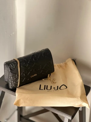 Svart kuvertväska från Liu Jo - Snygg svart kuvertväska från Liu Jo med präglat mönster och guldfärgad kedja. Väskan har elegant struktur och logga i guld framtill. Perfekt accessoar för att lyfta din outfit. Kommer med original dustbag.