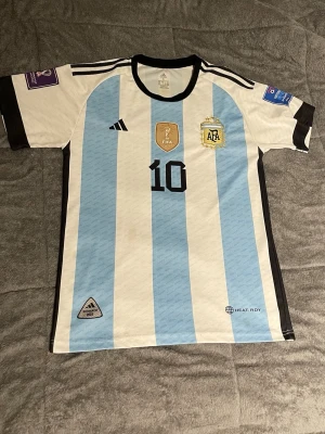 Argentina Messi matchtröja Adidas - Säljer en officiell Argentina matchtröja från Adidas med Messi och nummer 10 på ryggen. Tröjan har klassiska blå och vita ränder, svarta detaljer och FIFA-märke på bröstet. Tillverkad i lätt och ventilerande material, perfekt för fotboll eller samling.