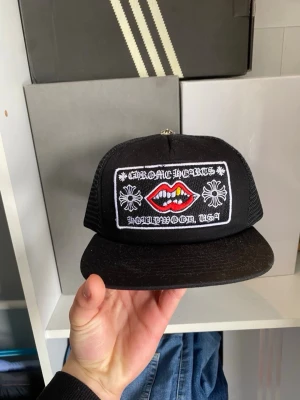 Svart Chrome Hearts truckerkeps - Säljer en svart truckerkeps från Chrome Hearts med mesh på sidorna och ett stort broderat patch framtill. Patchen har vita blommor, text och röda läppar med guldtänder. Justerbar snapback baktill. Perfekt för dig som gillar streetwear och statement-plagg.