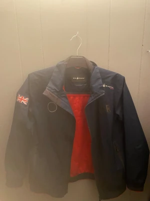 Marinblå vindjacka Sail Racing - Snygg marinblå vindjacka från Sail Racing med röd meshfoder och Union Jack-flagga på ärmen. Jackan har dragkedja, hög krage och logga på bröstet. Perfekt för dig som gillar sportig stil och vill ha en lätt jacka till blåsiga dagar.