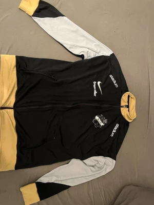 AIK träningtröja - Snygg AIK träningsjacka från Nike i svart med gula muddar och krage samt vita detaljer på ärmarna. Jackan har dragkedja, fickor och AIK:s klubbmärke på bröstet. Sponsorer som Stadium, Truecaller, SVEA och Fabege tryckta på fram- och baksidan. Perfekt för träning eller match.