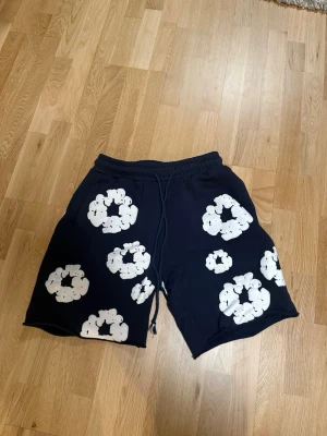 Svarta shorts med vita blommor - Säljer ett par svarta shorts med vita blommönster och snörning i midjan. Shortsen har en avslappnad passform och elastisk midja för extra komfort. Perfekta för sommardagar och har fickor både fram och bak.