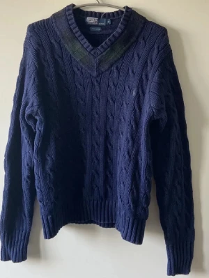 Mörkblå kabelstickad tröja Polo - Mörkblå kabelstickad tröja från Polo by Ralph Lauren i 100% bomull. Tröjan har V-ringning med ribbstickade detaljer och långa ärmar. Klassisk och tidlös design!                                                  Byten kan vara intressant!