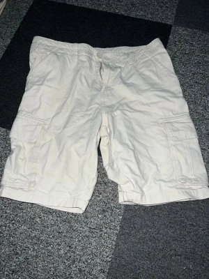 Beiga cargoshorts från 157 - Säljer ett par beiga cargoshorts från 157 i storlek S. Shortsen har flera praktiska fickor på sidorna och klassisk cargomodell. Perfekta för sommaren och riktigt sköna att bära till t-shirt eller hoodie.