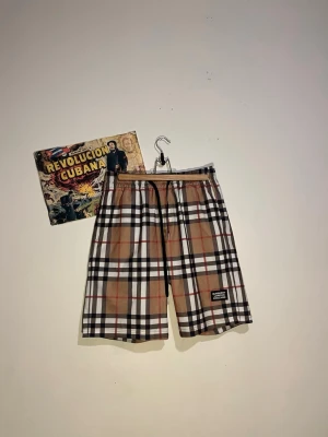 Rutiga shorts från Burberry - Säljer ett par klassiska rutiga shorts från Burberry med elastisk midja och snörning. Shortsen har det ikoniska beige, svarta, vita och röda mönstret samt en patch med Burberry London England på benet. Perfekta för sommaren och riktigt snygga till streetwear.