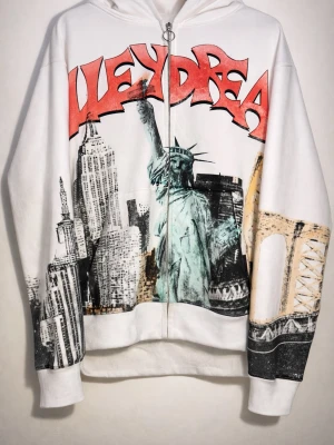 Vit hoodie med New York-print - Vit hoodie med dragkedja och stor huva. Framsidan har ett färgstarkt New York-motiv med Frihetsgudinnan, skyskrapor och broar samt röd graffiti-text. Oversized passform och ribbade muddar. Perfekt för dig som gillar streetwear och unika tryck.