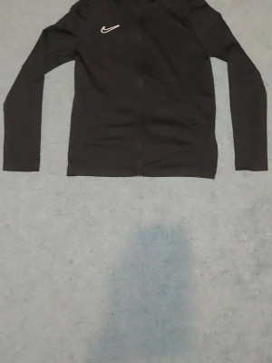 Svart Nike zip-tröja - Säljer en svart zip-tröja från Nike med lång ärm och vit Nike-logga på bröstet. Tröjan har en smal passform och är tillverkad i ett mjukt, stretchigt material som passar perfekt till träning eller chill. Dragkedja framtill och enkel design.