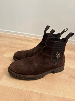 Mörkbruna Chelsea boots i mocka - Säljer ett par mörkbruna Chelsea boots från Polo Ralph Lauren i mjuk mocka. Skorna är sköna att ha på sig, dragflikar fram och bak samt en broderad logga i guld på sidan. Perfekta för dig som vill ha en stilren och klassisk look.
