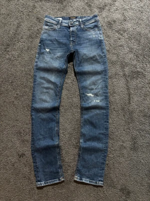 Jack & Jones jeans - Säljer ett par blå skinny jeans med slitningar och distressed detaljer på benen. Jeansen har klassisk femficksdesign, normal midja och dragkedjegylf. Perfekta för en avslappnad och trendig look.