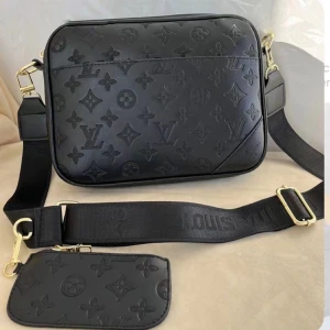 Svãrt axelväska 3 delar  - Snygg svart trio bag med klassiskt monogrammönster i präglat läder. Väskan har bred justerbar axelrem i textil med guldfärgade metalldetaljer och en matchande liten avtagbar väska. Perfekt för dig som gillar lyxig och stilren design.