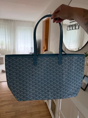 Blå väska från Goyard - Snygg väska från Goyard i blått med det klassiska Goyard-mönstret. Väskan har två långa handtag och är tillverkad i canvas med läderdetaljer. Perfekt storlek för att få plats med allt du behöver. En riktig statement piece för dig som gillar exklusiva accessoarer.
