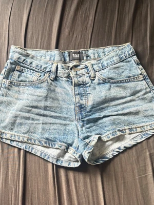 Blå denimshorts från 157 med brodyr - Säljer ett par klassiska blå denimshorts från 157 med hög midja och snygg broderad detalj på bakfickorna. Shortsen har fem fickor, bälteshällor och stängs med knapp och dragkedja. Perfekta till sommaren och ger en avslappnad vibe.
