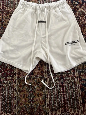 Essentials Fear of God - Snygga vita shorts från Essentials Fear of God med tryckt logga på benet och långa snören i midjan. De har en relaxed fit och elastisk midja för extra komfort. Perfekta för en chill och trendig look.