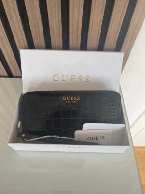 Svart väska krokopräglat skinn Guess - Snygg svart plånbok från Guess i krokopräglat konstläder med guldfärgade detaljer och dragkedja runtom. Plånboken har Guess-logga i guld framtill och medföljande handledsrem. Perfekt för dig som vill ha en stilren och trendig accessoar. Äkta
