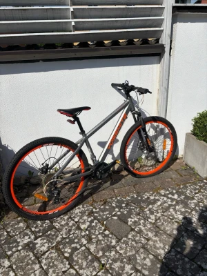 Grå och orange mountainbike - Säljer en grå mountainbike från Rockrider med orange detaljer och svart sadel med orange inslag. Cykeln har grova däck, svart styre och en kul ringklocka formad som en fotboll. Perfekt för dig som gillar att cykla i terräng eller på stan.