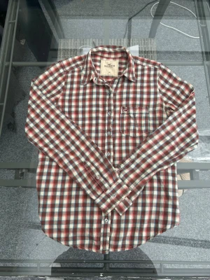 Hollister Rutig Flanellskjorta – M - Säljer en klassisk och skön flanellskjorta från Hollister i storlek M. Skjortan har en tidlös rutig design i varma toner av rött och brunt, vilket gör den perfekt för en avslappnad vardagsstil eller som lager-på-lager under hösten. Den är tillverkad i 100% bomull vilket gör den väldigt mjuk och bekväm att bära. Skjortan har en snygg bröstficka med broderad logga och är i fint skick. Kort info:  Märke: Hollister  Storlek: M  Material: 100% bomull  Skick: Fint använt skick