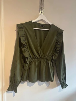 Olivgrön volangblus från Boohoo - Snygg olivgrön blus från Boohoo med djup V-ringning och volangdetaljer längs axlarna. Blusen har långa ärmar med resår vid ärmslut och markerad midja för en feminin siluett. Perfekt att styla med jeans eller kjol.