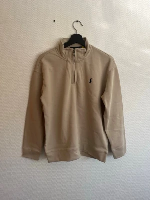 Beige halfzip från Polo Ralph Lauren - Snygg beige halfzip-tröja från Polo Ralph Lauren med klassisk broderad logga på bröstet. Tröjan har hög krage med dragkedja och är tillverkad i mjukt bomullsmaterial. Perfekt för en clean och stilren look. (obs ej orginal) 