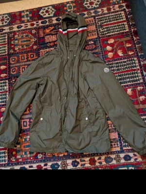 Olivgrön Moncler vindjacka - Snygg olivgrön vindjacka från Moncler med huva och dragsko. Jackan har två fickor med tryckknappar och Moncler-logga på ärmen. Materialet är lätt och vattenavvisande, perfekt för blåsiga dagar. Huva med röd och vit detalj upptill.