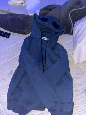 Blå hoodie med glasögon från C.P. Company - Säljer en blå hoodie från C.P. Company med ikoniska glasögon i huvan och dragkedja framtill. Hoodien har ribbade muddar och fickor på sidorna. 