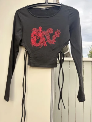 Svart croppad topp med röd drake  - Trendig svart croppad topp från SHEIN med lång ärm och coolt rött drakmotiv på bröstet. Toppen har snörning i sidorna för en justerbar passform och är tillverkad i mjuk bomullsmix. Perfekt för dig som vill sticka ut!storlek 36 men kan även passa xs.