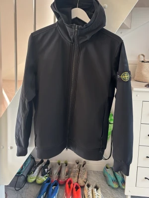 Svart vindjacka från Stone Island - Snygg svart vindjacka från Stone Island med huva och dragkedja framtill. Jackan har den klassiska logotypen på ärmen och en minimalistisk design. Perfekt för dig som gillar streetwear och vill ha en stilren look