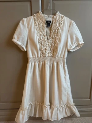 Klänning med volang från H&M - Superfin crèmefärgad klänning från H&M med volangdetaljer med lyxig känsla i tyget. Mycket bra skick!