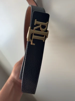 Svart bälte från Ralph lauren 120cm - Fint svart bälte med guldigt spänne. Brun färg på insidan om hellre vill ändra färgen på bältet. 120cm. Använd 1-2 gånger. Dock fått en mindre repa på spännet- se bild 