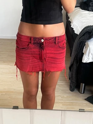 Röd jeanskjol med rå kant - Säljer en kort röd jeanskjol med rå, fransig kant och klassiska fem fickor. Kjolen har bälteshällor och dragkedja framtill. Perfekt för dig som gillar en edgy och trendig stil. Midjemått rakt över: 36cm. Längd: 26cm 