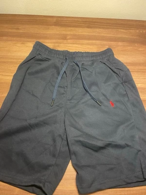 Svarta shorts från Polo Ralph Lauren - Svarta shorts från Polo Ralph Lauren med elastisk midja och snörning. Klassisk röd logga broderad på vänster ben. Tillverkade i mjukt material, perfekta för sommardagar. Två sidofickor för enkel förvaring.
