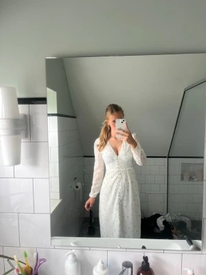 Vit långklänning med blå blommor - Romantisk vit långklänning med små blå blommor, v-ringning och knappar framtill. Klänningen har långa, transparenta ärmar och markerad midja för en feminin siluett. Perfekt för sommarens alla tillfällen. Endast använd 2x, säljs då den ej kommer till användning längre
