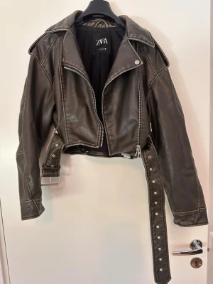 Svart skinnjacka från Zara - Snygg svart skinnjacka från Zara med klassisk bikerstil, bred krage och silvriga dragkedjor. Jackan har ett bälte i midjan och detaljerade sömmar på axlar och ärmar. Perfekt för dig som vill ha en edgy look.