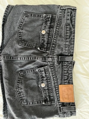 Mini-jeanskjol från Levi's - Säljer en svart/grå jeanskjol från Levi's i storlek XS. Kjolen har klassiska fem fickor, bälteshällor och en snygg läderpatch bak. Gjord i slitstark bomull med en kort och rak passform. Perfekt till sneakers eller boots.