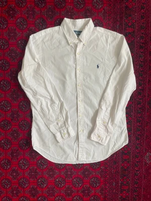 Ralph Lauren Skjorta - Klassisk vit skjorta från Ralph Lauren med broderad marinblå logga på bröstet. Skjortan har lång ärm, knappar framtill och krage. Tillverkad i bomull för en skön och tidlös look.