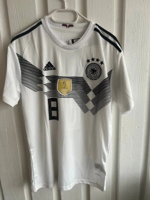Tysklands fotbollströja Kroos #8 Adidas - Vit officiell Tyskland fotbollströja från Adidas med svarta detaljer och tryck. Nummer 8 och Kroos på ryggen, samt FIFA-märke och landslagets emblem på bröstet. Korta ärmar, klassisk rund hals och tillverkad i lätt funktionsmaterial.