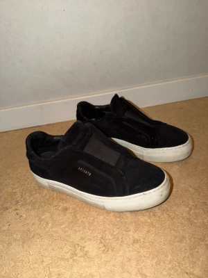 Svarta sneakers från Axel Arigato - Stilrena svarta sneakers från Axel Arigato i mocka med vit sula och elastiskt band över vristen istället för snören. Diskret logga i guld på sidan. Perfekta för en clean och modern look.