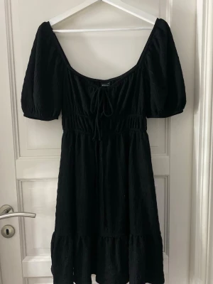 Svart kort klänning med puffärm - Trendig svart kort klänning med puffärmar och djup urringning. Klänningen har resår i midjan, knytning framtill och volang nedtill. Perfekt för dig som gillar en feminin och modern stil.