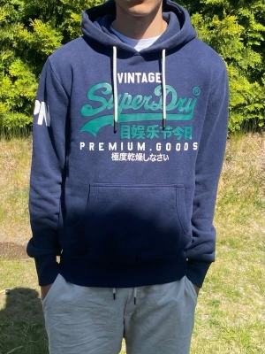 Mörkblå Superdry hoodie med tryck - Mörkblå hoodie från Superdry med stort turkost och vitt tryck på bröstet samt japanska tecken. Klassisk känguruficka, vita dragsnören och mjukt fleecefoder på insidan. Superdry-logga även på ärmen. Perfekt för en avslappnad streetwear-look.