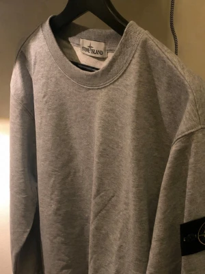 Stone Island hoodie - Säljer en stilren grå sweatshirt från Stone Island med klassisk rund halsringning och den ikoniska logotypen på ärmen. Tröjan är i mjukt bomullsmaterial och har en avslappnad passform som passar perfekt till streetwear-stilen.