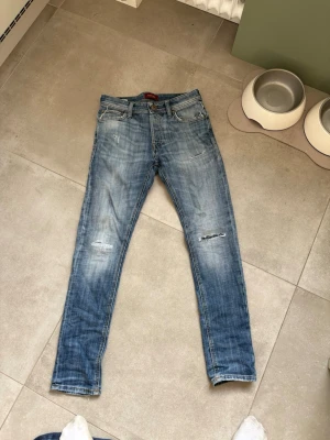 Jack & Jones jeans med slitningar  - Säljer ett par ljusblå jeans från Jack & Jones med slitningar och distressed detaljer på benen. Modellen är slim fit med klassisk femficksdesign och knappgylf. Perfekta för en avslappnad och trendig look.