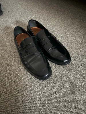 Svarta klassiska loafers i skinn - Säljer ett par stilrena svarta loafers i blankt skinn med bruna innersulor. Skorna har en tidlös design med rundad tå och diskret söm längs ovansidan. Perfekta för dig som gillar en clean och klassisk look.
