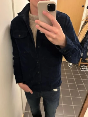 Mörkblå overshirt från Jack & Jones - Snygg mörkblå overshirt från Jack & Jones i manchestertyg. Klassisk krage, knappstängning framtill och bröstficka med lock. Perfekt lager på lager-plagg för en avslappnad stil.