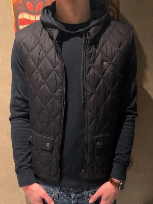 Svart dunväst - Säljer en svart quiltad dunväst med dragkedja och två fickor med tryckknappar. Västen har ett snyggt rutmönster och är perfekt att slänga över en hoodie eller tshirt . Materialet är lätt och isolerande, vilket gör den både bekväm och stilren, samt varm. Modellen är 185cm